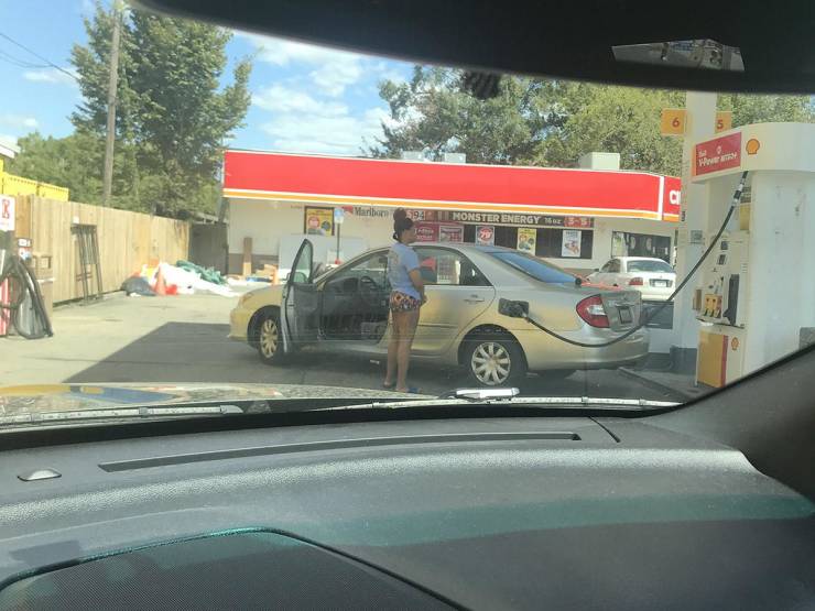 Žena pri čerpacej stanici tankuje auto a kontroluje čerpadlo. V pozadí obchod s červenou strechou.