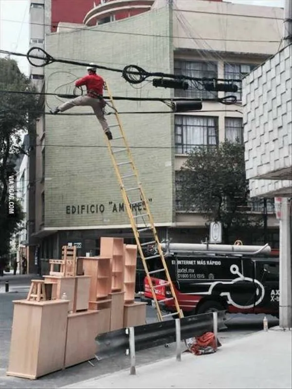 Pracovník sa snaží dosiahnuť káble cez vysoký rebrík, ktorý je podoprený nestabilnou stoličkovou konštrukciou na ulici.