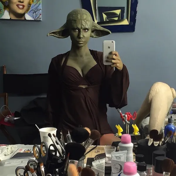 Osoba s maskou sci-fi postavy pózuje pred zrkadlom v maske a kostýme zeleného mimozemšťana, okolo sú líčidlá.