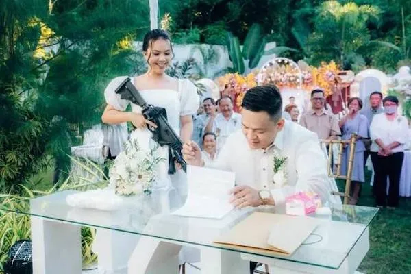 Nevesta s úsmevom drží repliku zbrane, zatiaľ čo ženích podpisuje dokument pred svedkami na svadobnom obrade.