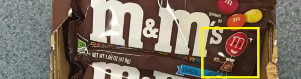 Obal sladkostí M&M's s logom a farebnými cukríkmi na tmavom pozadí, zdôraznený obrázok maskota v pravom dolnom rohu.