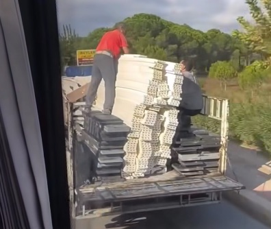 Muž odváža stavebný materiál na korbe auta po ceste, na okolitom pozadí sú stromy a zeleň.