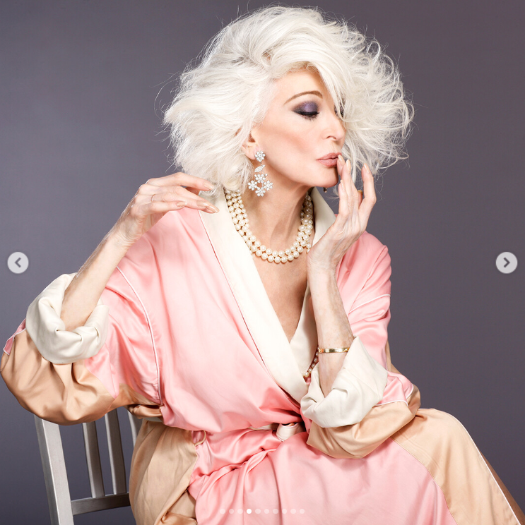 Elegantná žena s bielymi vlasmi pózuje v ružovom župane a perlových šperkoch proti tmavému pozadiu.
