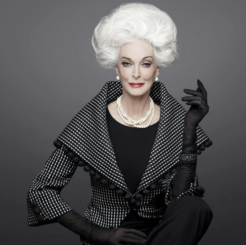 Staršia elegantná žena v čierno-bielom kabáte s perlovým náhrdelníkom, pózuje na tmavom pozadí.