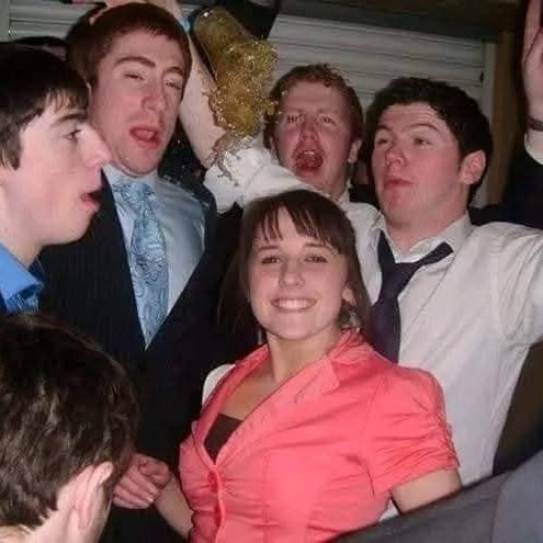 Skupina mladých ľudí na večierku s usmiatou ženou v popredí. Vzadu jeden muž drží nápoj.