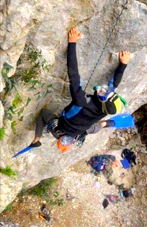 Horolezec v neopréne a plutvách lezie po strmej skalnej stene, kombinujúci potápanie a lezenie v nezvyčajnom štýle.