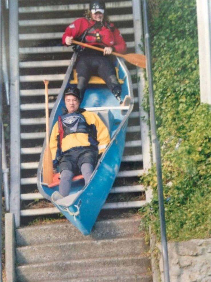 Kanoe na schodoch: Dobrodružná jazda dvoch mužov dolu schodmi v modrej kanoe na schodisku, s veslami v rukách.