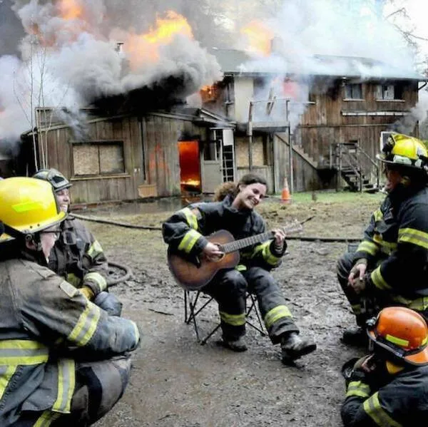 Hasiči sedia okolo kolegu hrajúceho na gitaru pred horiacim domom, kľudne sledujú situáciu.