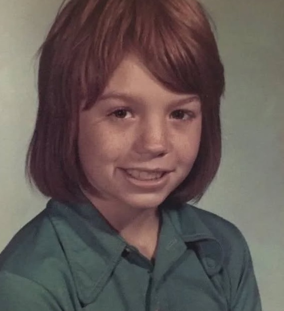 Osoba s hnedými vlasmi v tyrkysovej košeli sa usmieva na školskom fotografickom portréte.