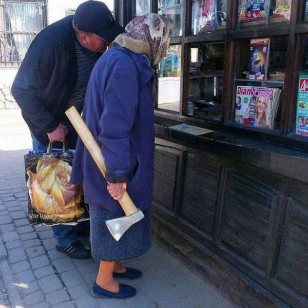 Staršia žena s motykou na ulici hľadí do výkladu s časopismi, vedľa nej stojí muž s nákupnou taškou.