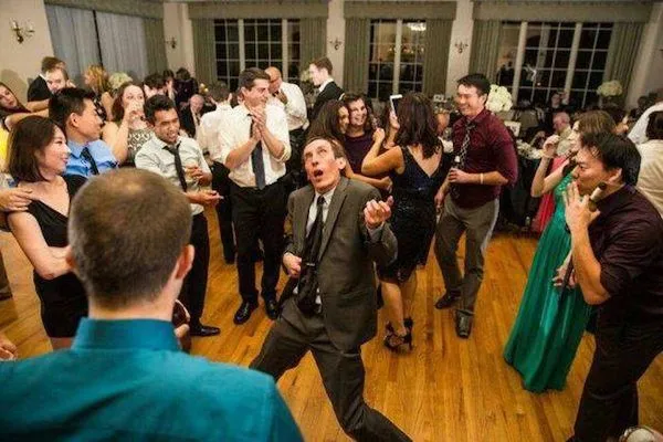 Skupina hostí tancuje a zabáva sa na večierku, uprostred je muž v obleku predvádzajúci extatické pohyby.