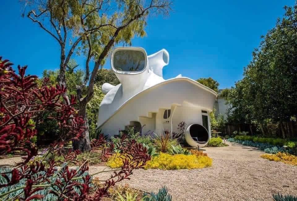 Moderný dom v tvare morskej mušle obklopený záhradou, s modrou oblohou v pozadí.
