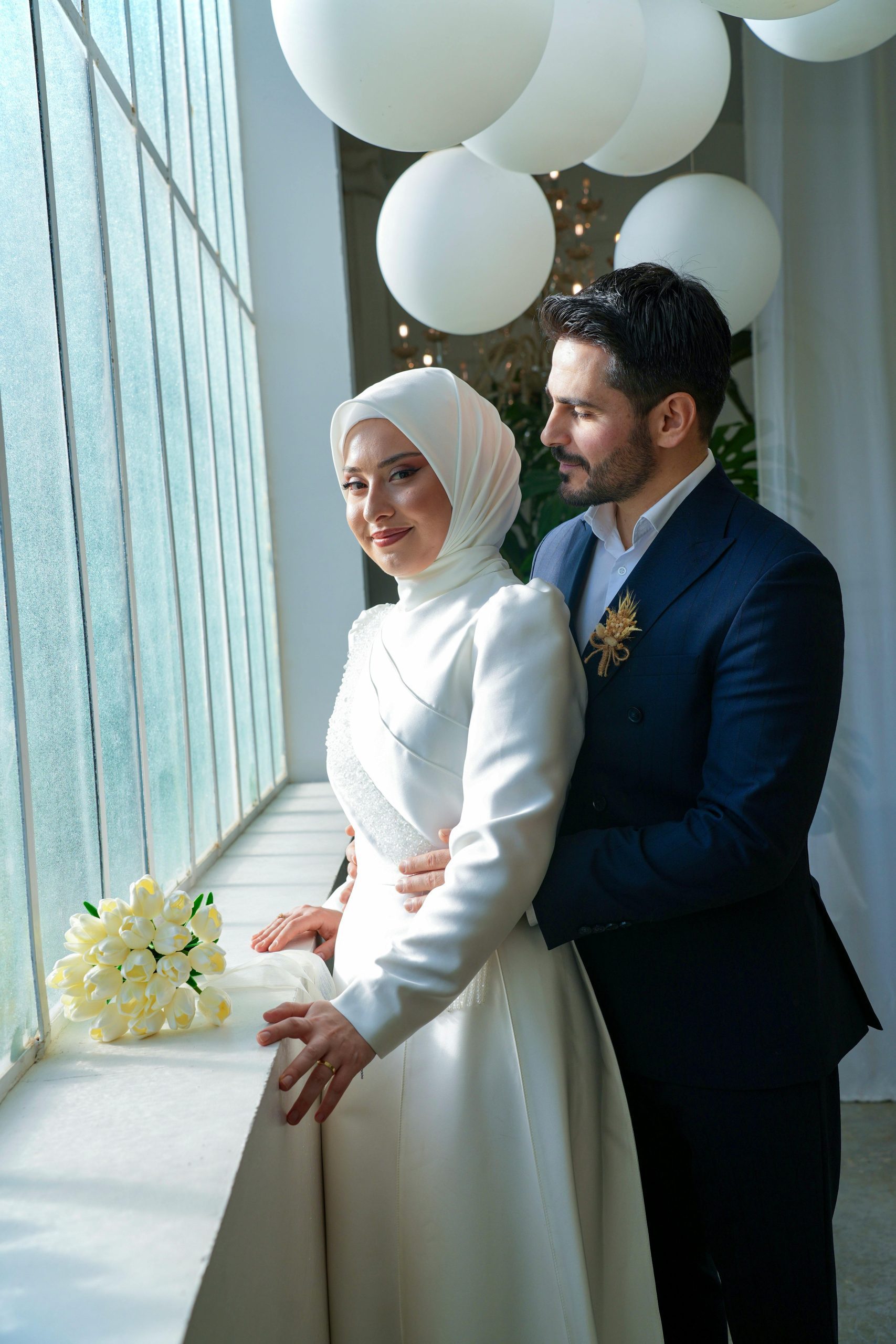 Pár oslavuje svadobný deň pri okne so slnečným svetlom, elegantné oblečenie, jemná kytica a balóny v pozadí.