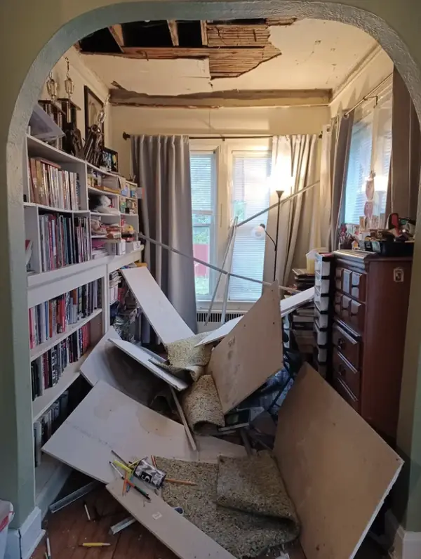Pokazený strop v obývačke s popadanými kusmi omietky, police s knihami a zatiahnuté závesy.