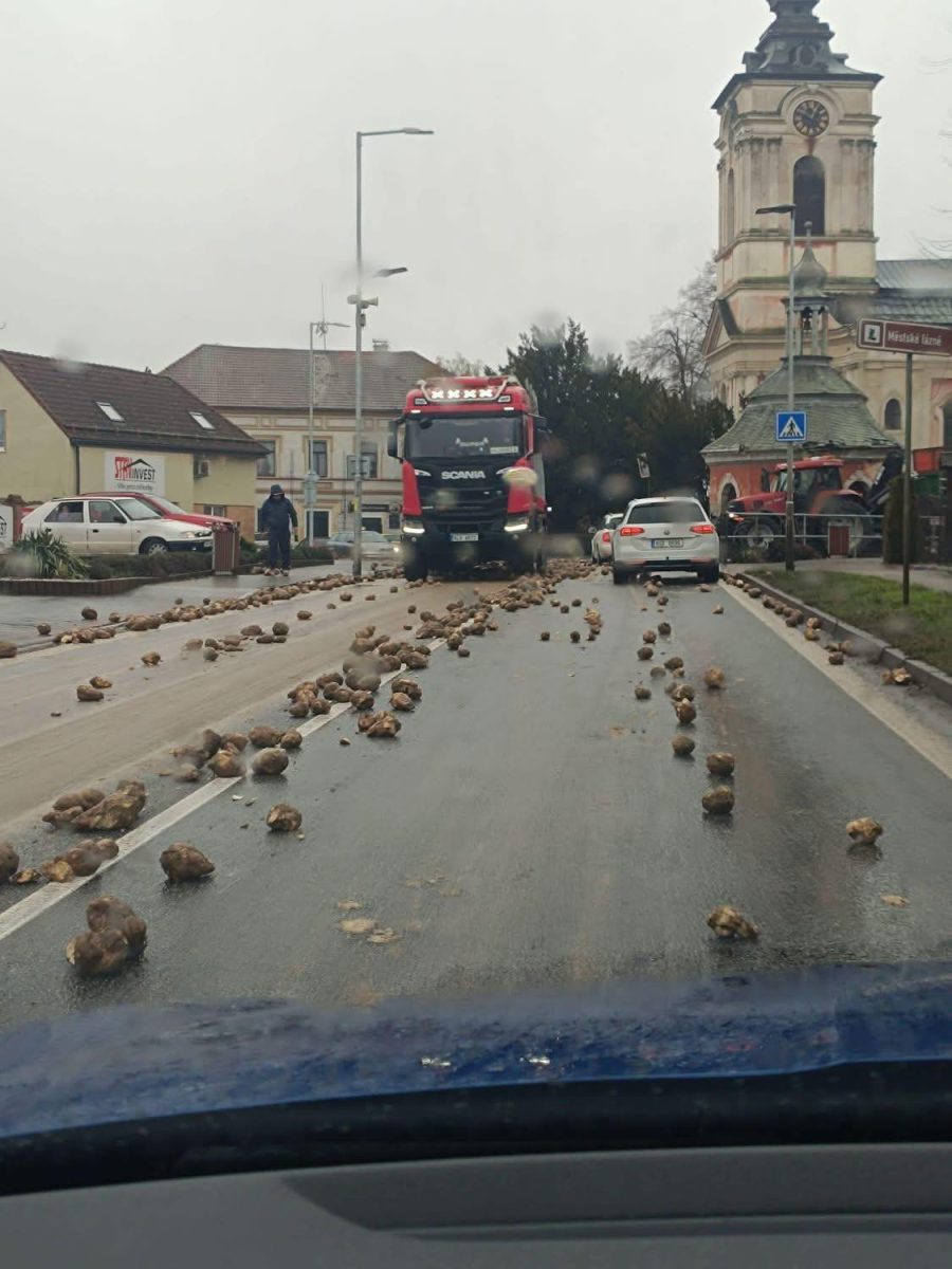 Dve autá obklopené roztrúsenými zemiakmi na ceste blízko centrálnej budovy v daždivom počasí.