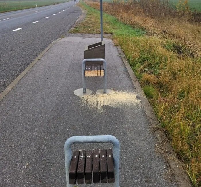 Lavičky na zastávke bez sedadiel, obklopené trávou a cestou, s nánosom drevených pilín na chodníku.