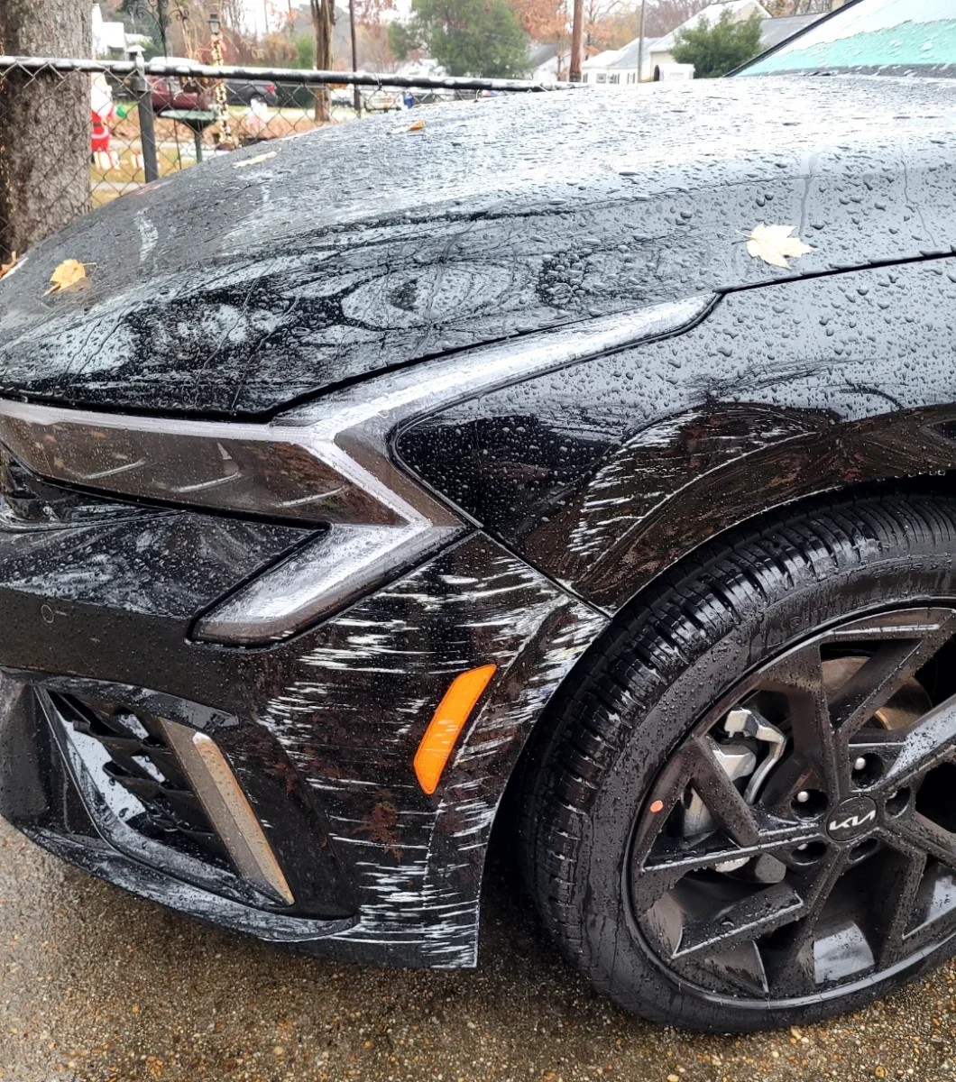 Čierne auto so škrabancami na prednom nárazníku po daždi, blízko kolesa s lesklými ráfikmi.