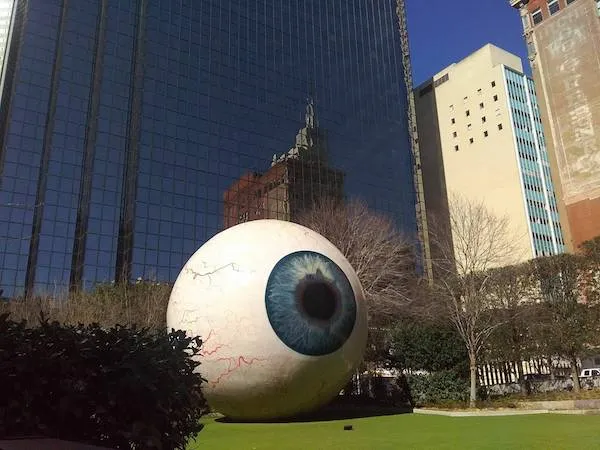 Obrovské umelecké dielo v tvare oka na trávniku pred modernou budovou v meste, za slnečného počasia.