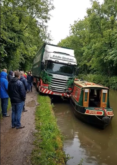 Kamión a loďka v úzkom prieplave pri ceste, skupina ľudí sleduje blízke stretnutie.
