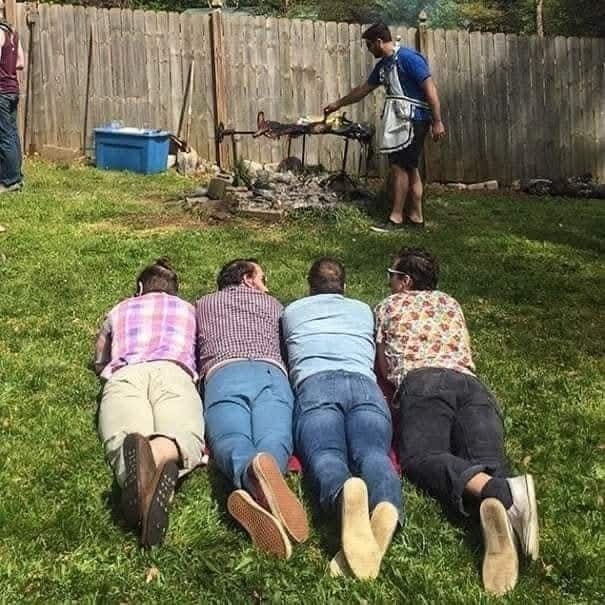 Skupina ľudí leží na tráve pred grilom, kde muž griluje. Letná grilovačka na záhrade.