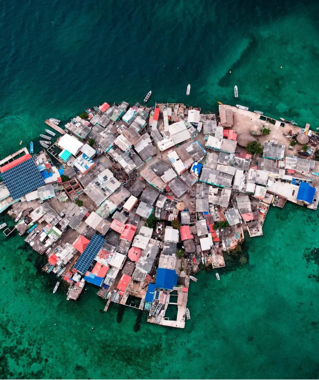 Letecký pohľad na hustú osadu obklopenú tyrkysovou vodou so strechami rôznych farieb.