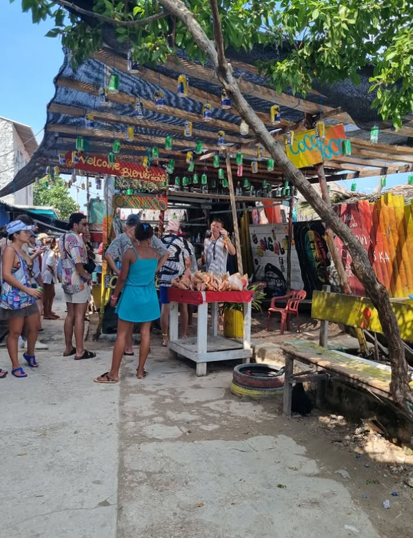 Trhovisko s ľuďmi, farebné stánky a nápis Welcome, tropická atmosféra pod stromami.