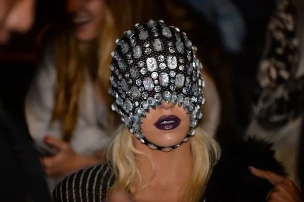 Osoba s extravagantným klobúkom pokrytým lesklými kamienkami, výrazný fialový rúž na perách.