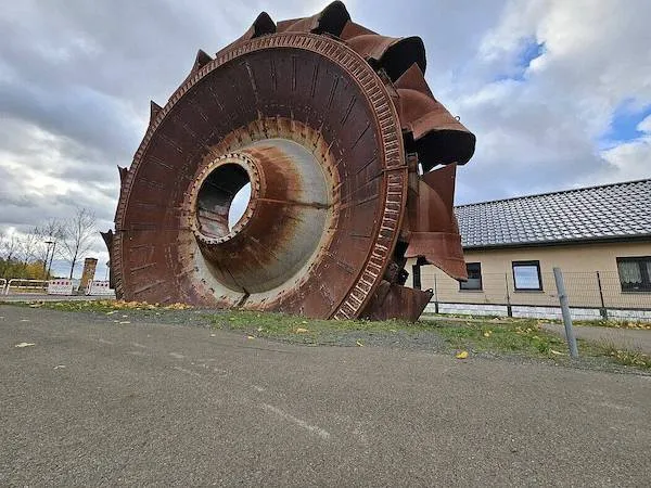 Starý banský stroj vystavený vonku, obloha je zamračená, v pozadí je dom so šikmou strechou.