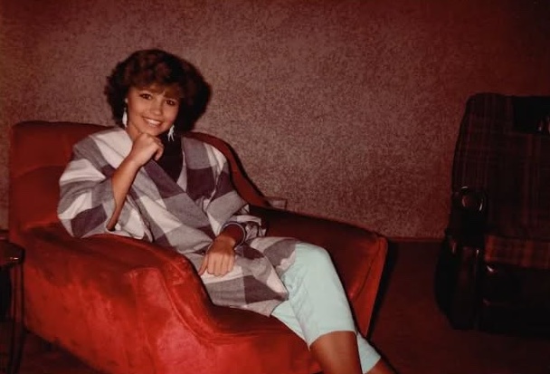 Osoba sedí v červenom kresle, oblečená v károvanej bunde a bledých nohaviciach, s úsmevom v interiéri.