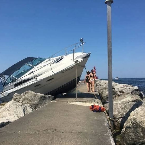 Loď narazená na mólo s kameňmi, dve ženy v plavkách, jasná obloha, dovolenková atmosféra.