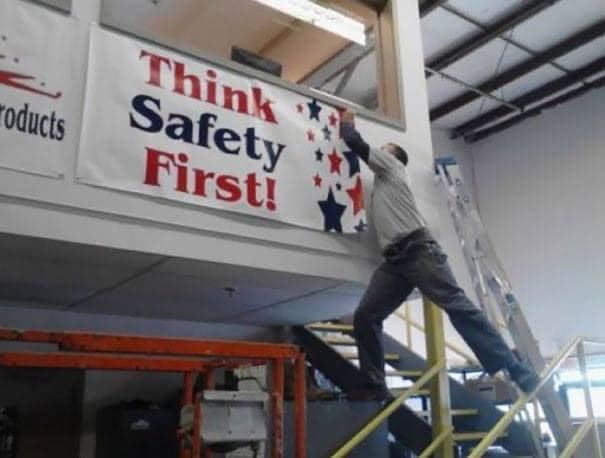 Muž na rebríku v interiéri inštaluje banner s nápisom Think Safety First!, ale stojí v nebezpečnej pozícii.