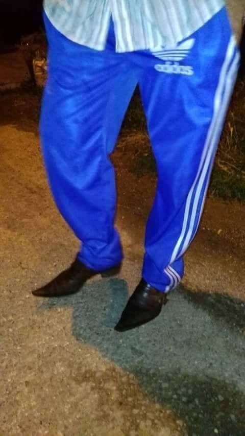 Muž v modrých tréningových nohaviciach s elegantnými topánkami kráča po ceste v noci.