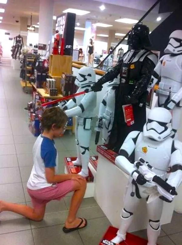 Dieťa kľačí pred figúrkami z Hviezdnych vojen v obchode, vrátane Darth Vadera a stormtroopera.