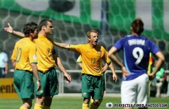 Futbalisti v žltých dresoch protestujú voči rozhodnutiu rozhodcu počas zápasu, zatiaľ čo súper stojí chrbtom.