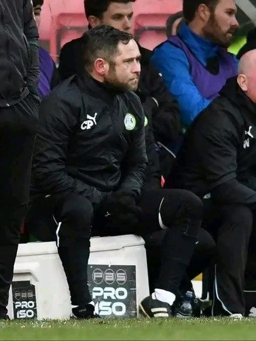 Tréner v teplom oblečení sedí na lavičke počas futbalového zápasu, sústredený na dianie na ihrisku.