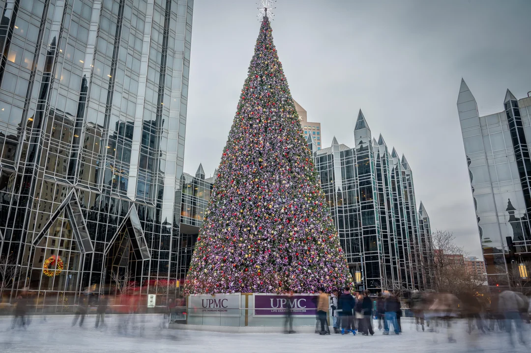 Vianočný stromček pred modernou budovou, obklopený ľuďmi na klzisku, zdobený pestrými svetlami a ozdobami.