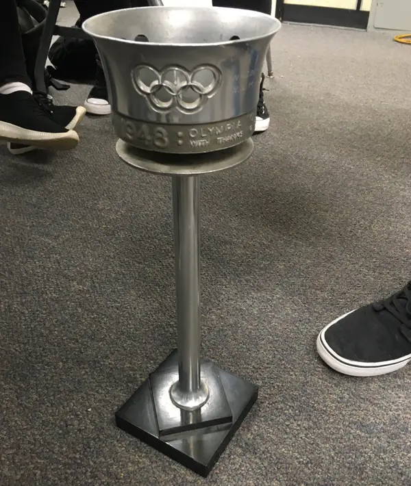 Olympijská fakľa z Londýna 1948 položená na podlahe, s viditeľným logom olympijských kruhov.