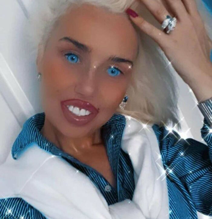 Neznáma osoba s blond vlasmi a intenzívnym úsmevom pózuje v modro-bielej košeli, s rukou vo vlasoch.