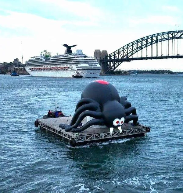 Nafukovací pavúk na vode pred ikonickým mostom a výletnou loďou v pozadí.