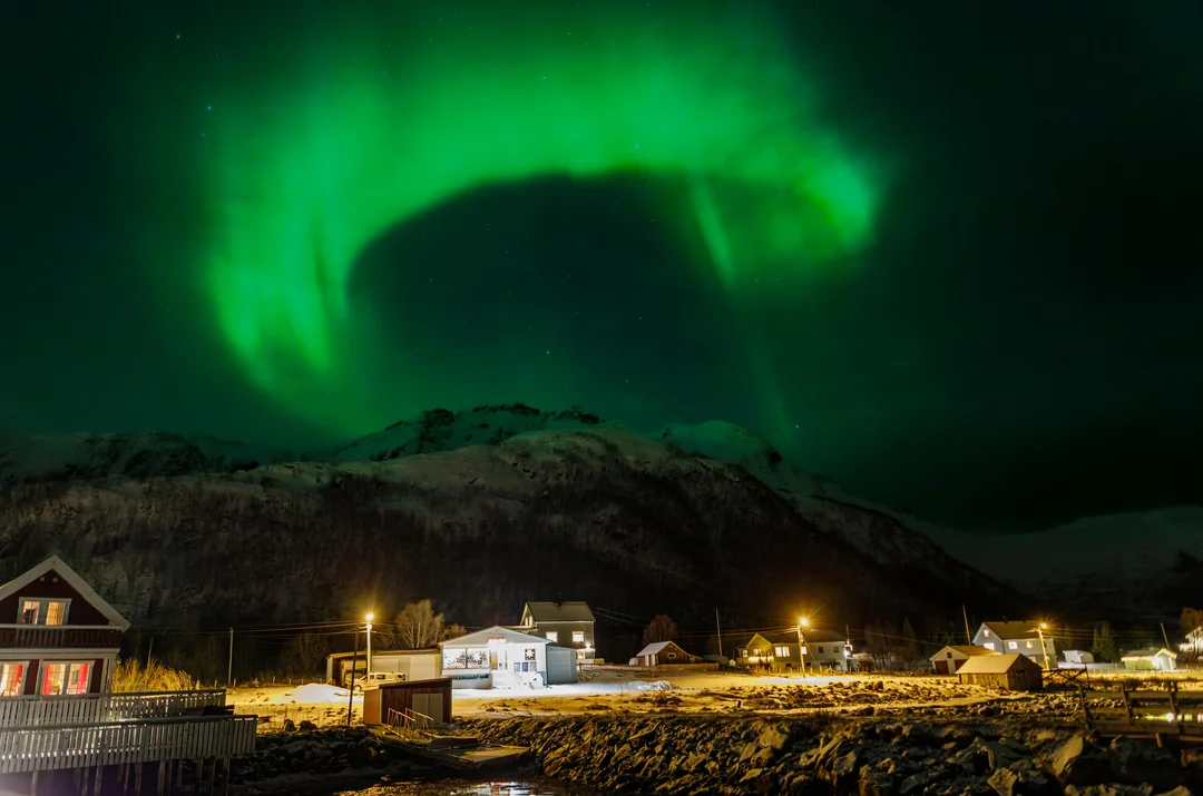 Polárna žiara tancuje nad zasneženou horskou dedinkou v noci, osvetlenie domov kontrastuje s tmavou oblohou.