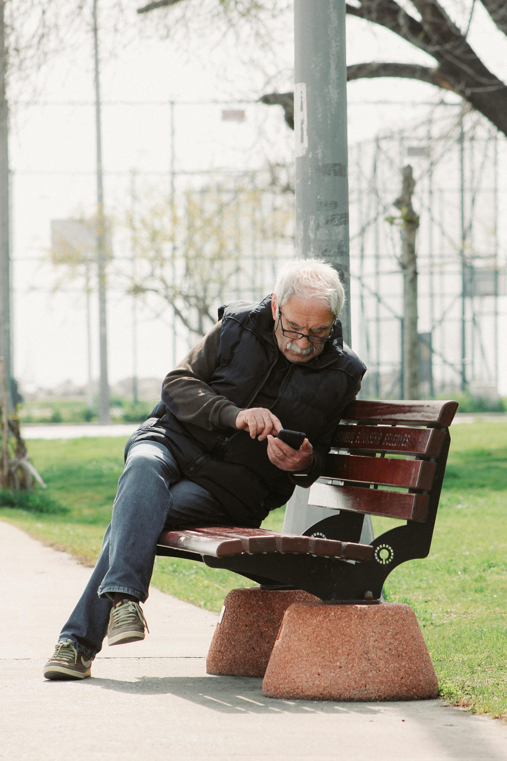 Starší muž v okuliaroch sedí na lavičke v parku a sústredene používa smartfón.