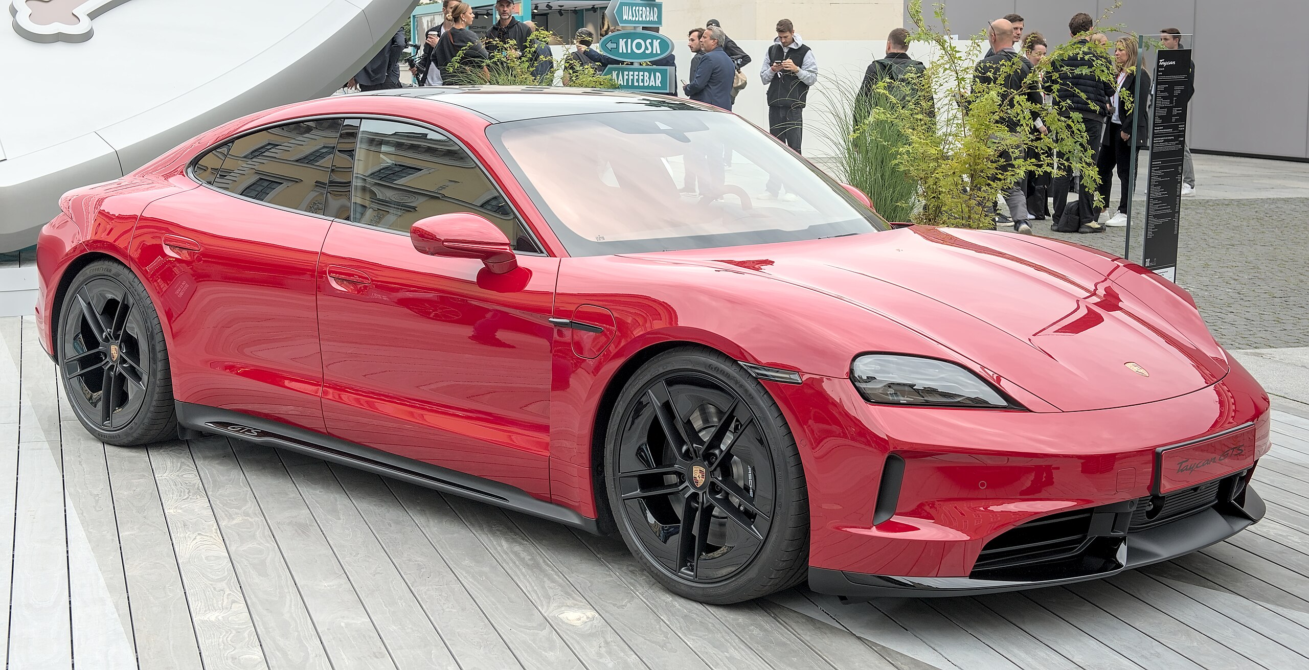 Červené športové auto zaparkované na drevenej podlahe s divákmi v pozadí.