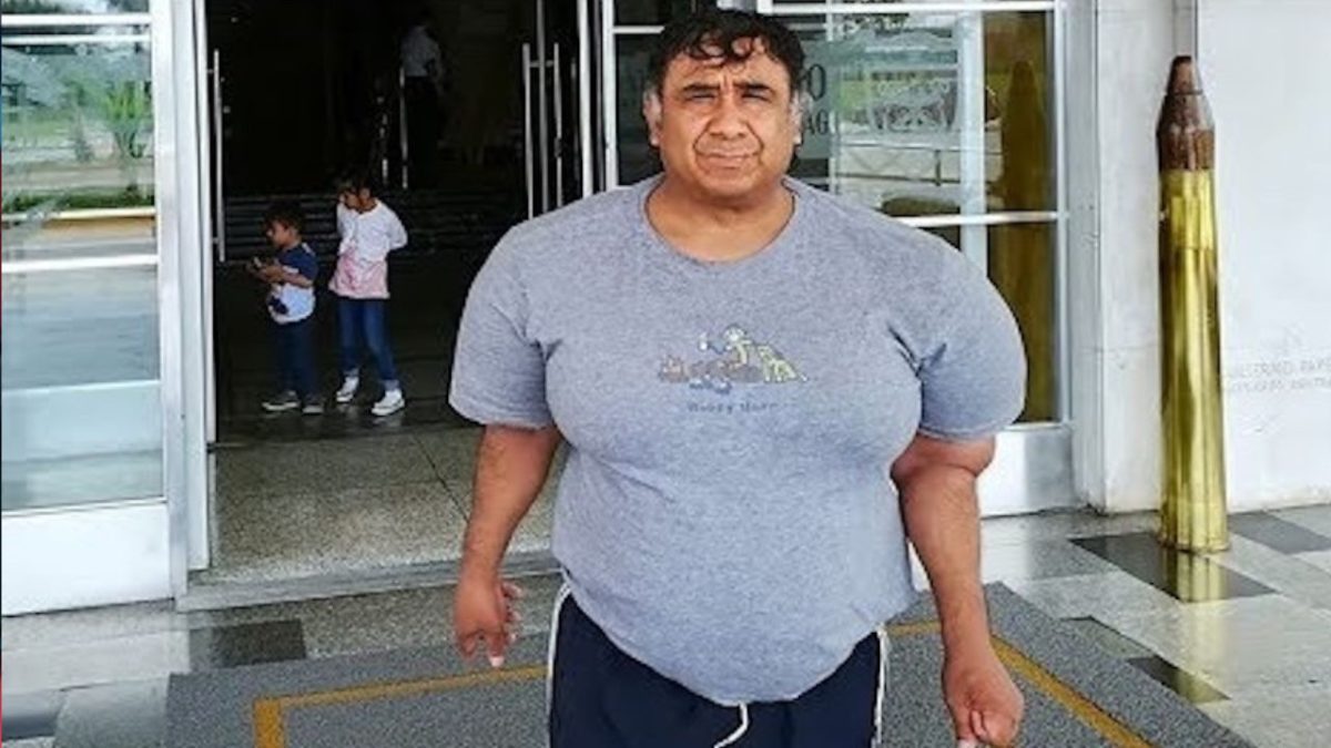 Osoba v šedej tričku stojí pred budovou. V pozadí sú viditeľné deti pri vstupe.