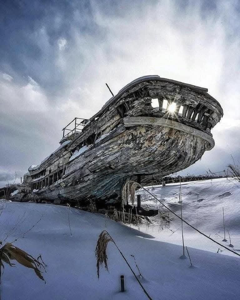 Starý vrak lode pokrytý snehom, slnečné lúče presvitajú cez konštrukciu na zimnej pláni.