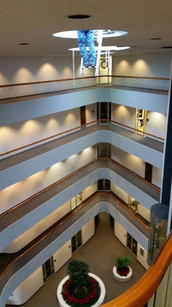 Pracovník na rebríku vykonáva opravy svetla v átriu viacpodlažnej budovy s moderným dizajnom.