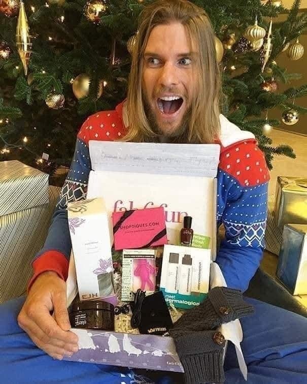 Muž s nadšením otvára vianočné darčekové krabice plné kozmetiky a doplnkov, sediac pri ozdobenom stromčeku.