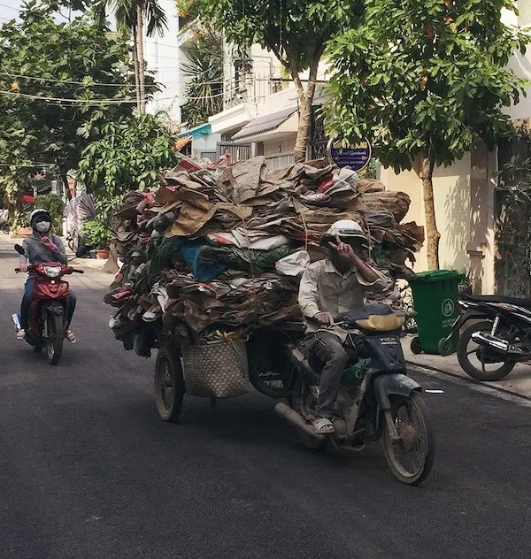Muž na motocykli vezie hromadu kartónov po ulici, vedľa prechádza ďalší motocykel.