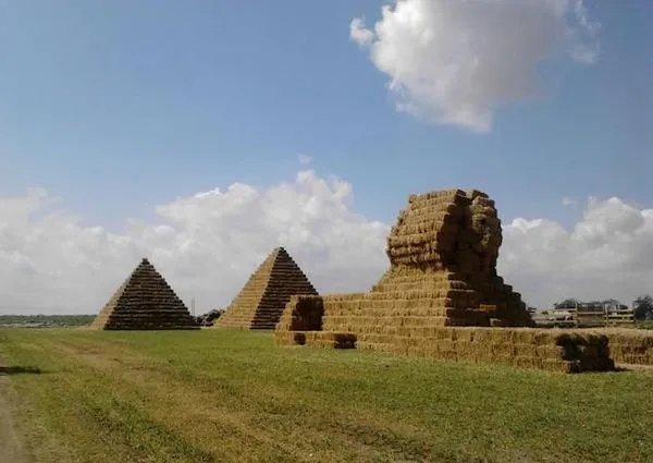 Slamené pyramídy a sfinga na poli pod modrou oblohou s oblakmi. Kreatívne štruktúry z balíkov slamy.