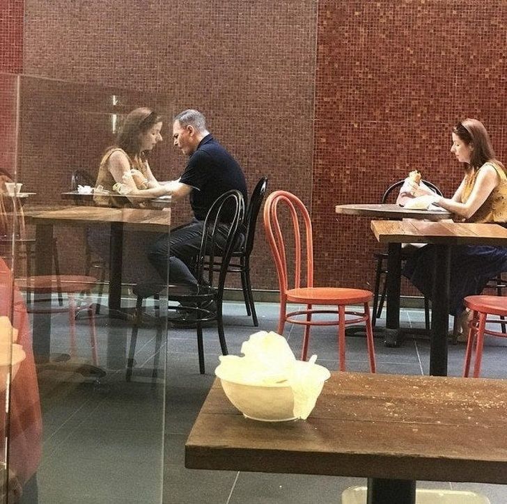 Dve osoby sediace v kaviarni pri stole, zrkadlenie odráža ich diskusiu. Vedľa nich žena jemne obeduje.