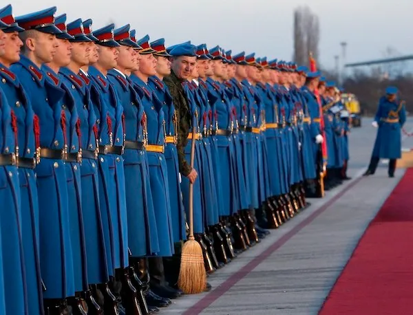 Skupina vojakov v modrých uniformách stojí na letisku v rade, jeden drží metlu, vedľa červeného koberca.
